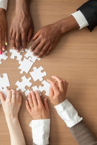 Hands of multi-ethnic team assembling jigsaw puzzle, multiracial group of black and white people joining pieces at desk, successful teamwork concept, help and support in business, close up top view